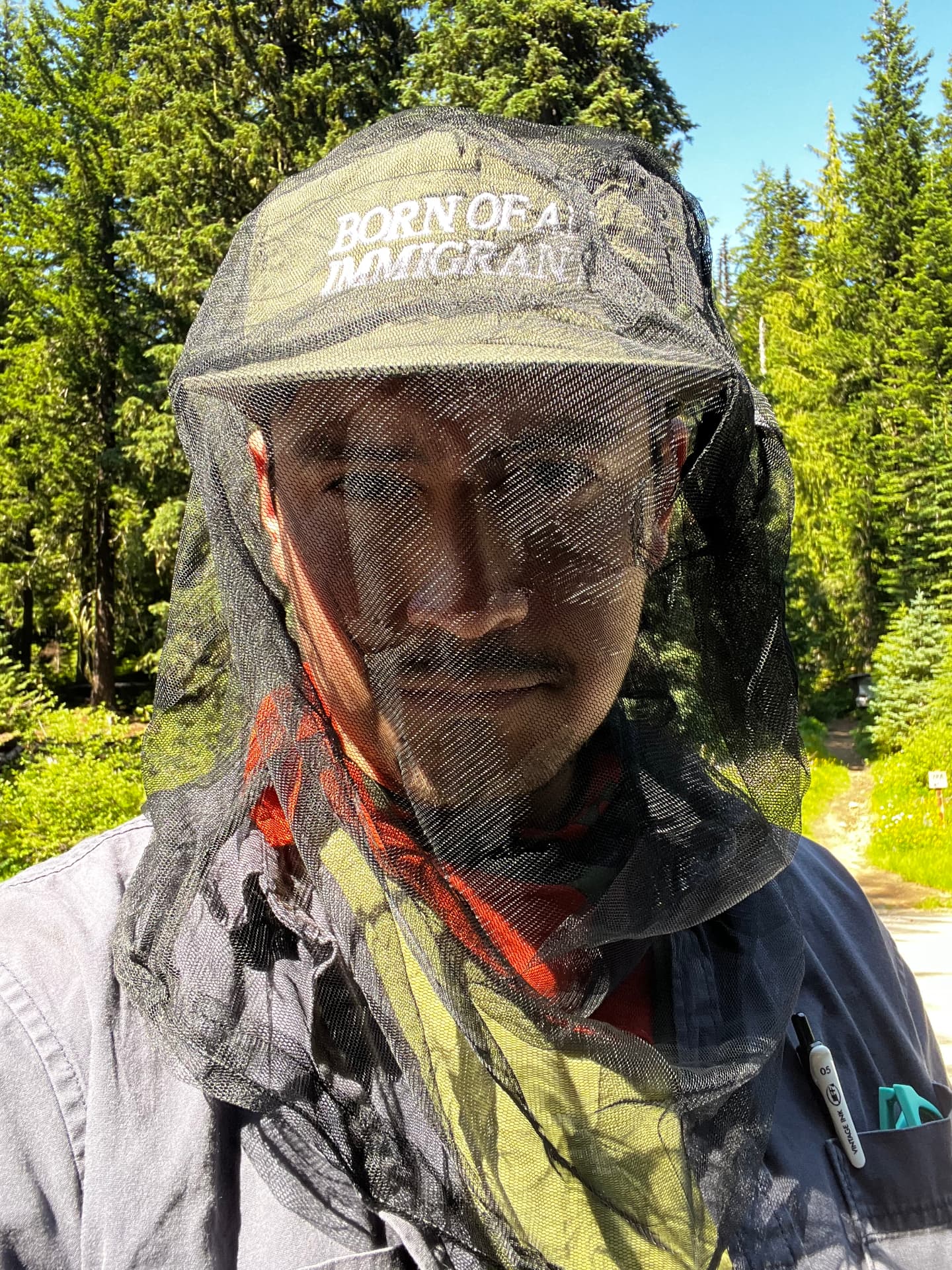 Wearing a mesh bug net over a BORN OF AN IMMIGRANT hat on a forest trail